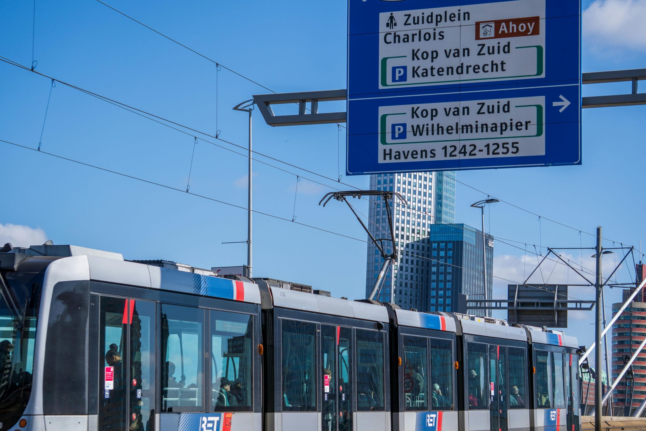 A modern tram passing by high-rise buildings in Rotterdam under a clear blue sky.